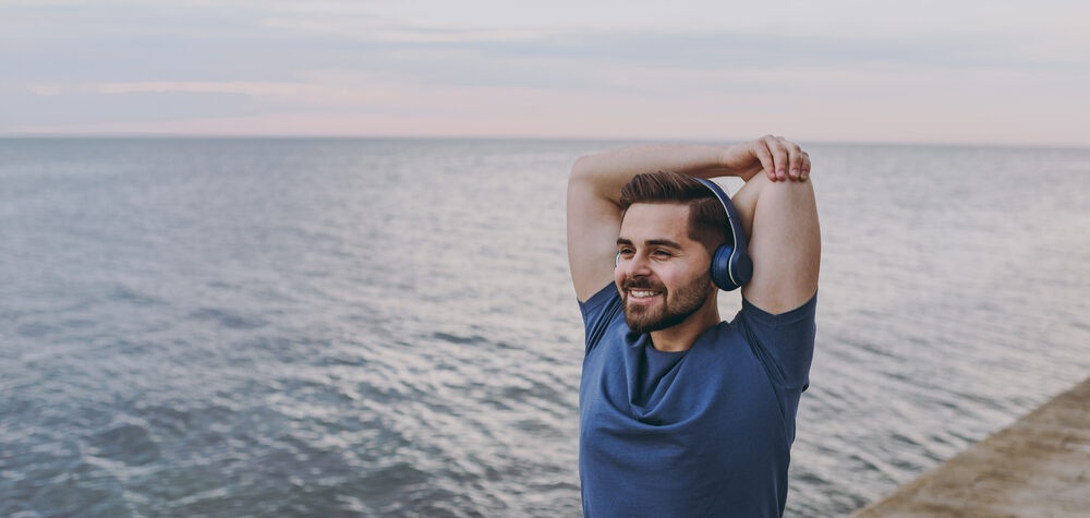 Man with brown hair smiles as he stretches along the beach before going for a run