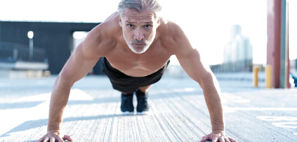 Middle aged man does pushups on concrete while looking directly into the camera