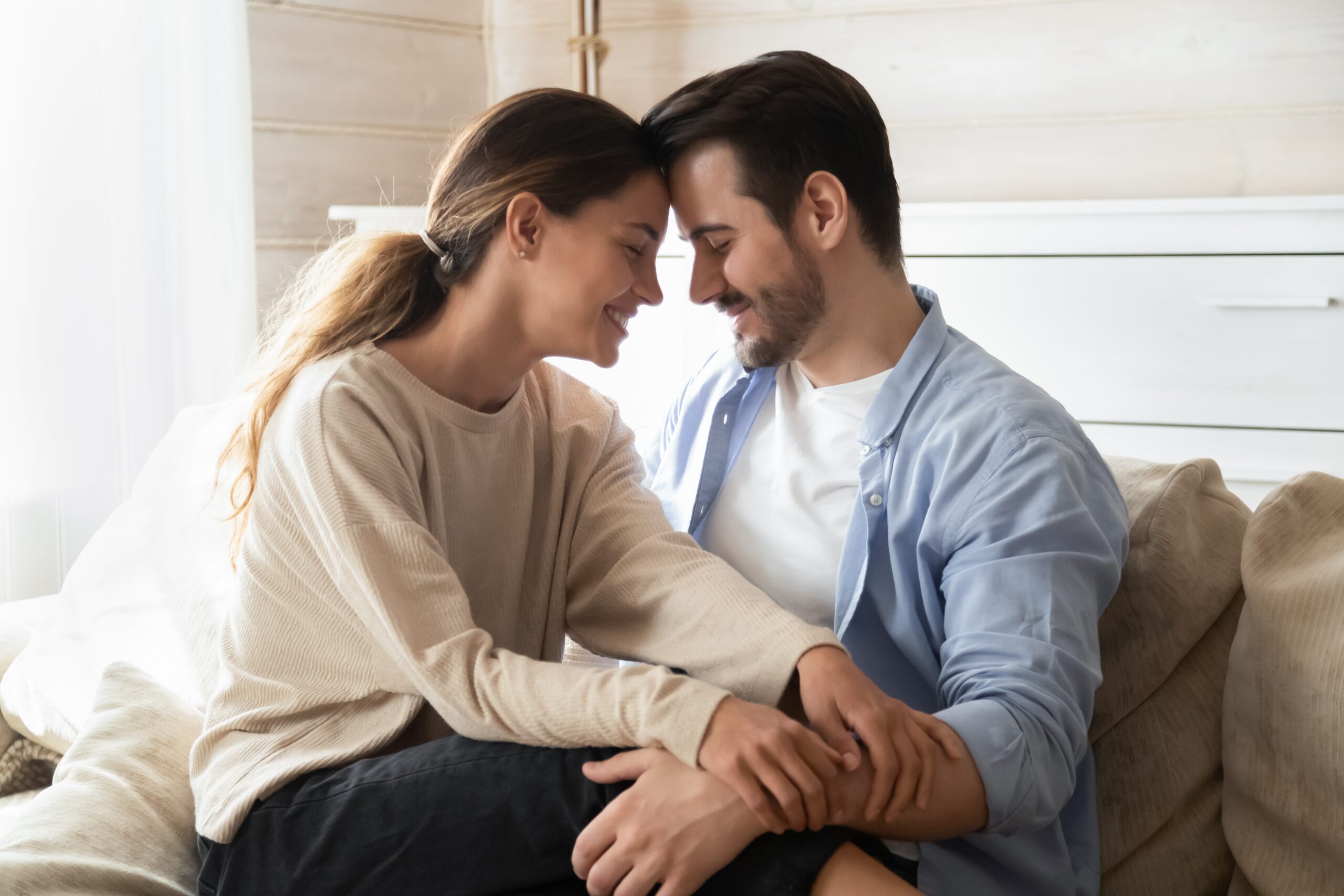 A couple embraces on the couch while smiling and putting their heads together.